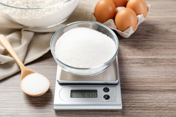 Digital kitchen scale with bowl of sugar and spoon on wooden table, closeup