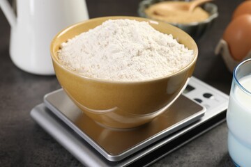Digital kitchen scale with bowl of flour and other products on grey table, closeup