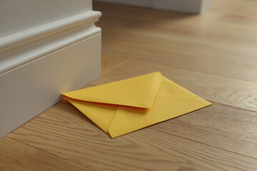 Yellow envelope lies on light brown wood floor near white baseboard, suggesting secrecy, mystery, or a hidden message
