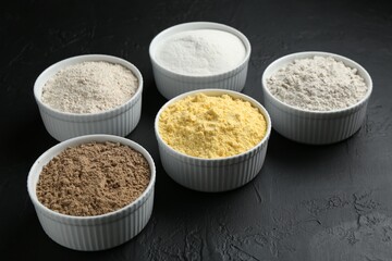 Different types of flour in bowls on black textured table, closeup