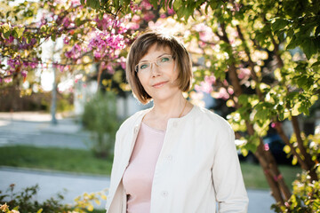 Smiling blonde mature woman 55-60 year old wearing eye glasses posing over flowers in park outdoors close up. Looking at camera. Wellness lifestyle.