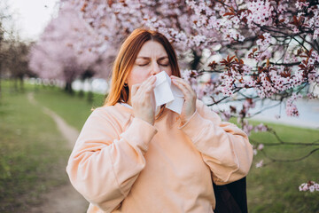 Sneezing young redhead woman with nose wiper among blooming trees in park. Portrait of sick women...
