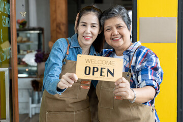 Two Asian female cafe owners wearing aprons smiling together while holding welcome open sign in front of shop entrance. Expressing teamwork, joy, support, and proud small business ownership.