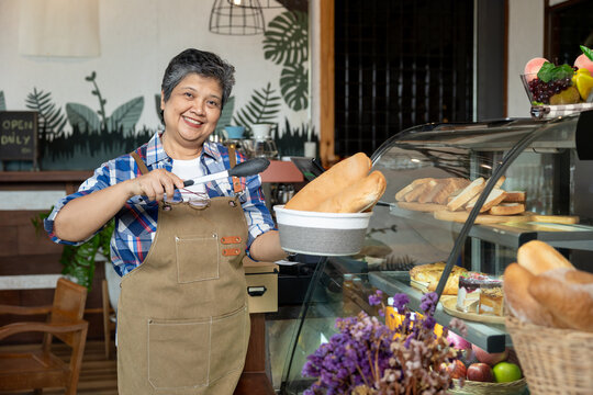 Asian senior woman wearing apron holding basket of bread using tongs to serve inside cozy cafe. Smiling with friendly face, representing local bakery owner working with joy and care in daily routine.
