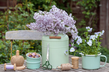 Garten-Arrangement mit Fliederstrauß, Hornveilchen im Topf und Garnrollen