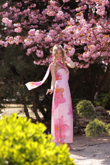 beautiful woman with blond hair in elegant dress and accessories posing in blooming sakura garden