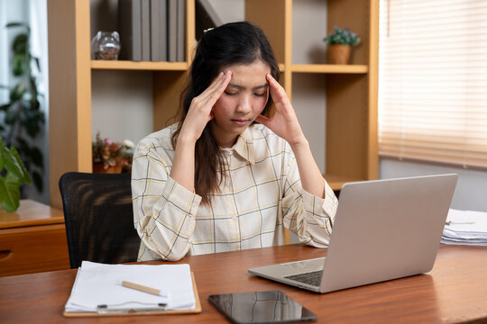 Young Asian female student sitting with eyes closed and holding head feeling headache and overwhelmed from online learning using laptop at home with tablet paper pen and shelf in background
