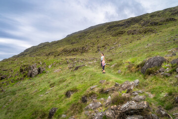 Hikers stands on a grassy hillside of Slieve Foye, surrounded by rocky terrain and vibrant lush greenery beneath a cloudy sky, enjoying a peaceful moment in nature, Carlingford, Ireland