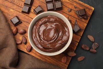 Tasty pudding in bowl, pieces of chocolate and cocoa beans on black table, flat lay