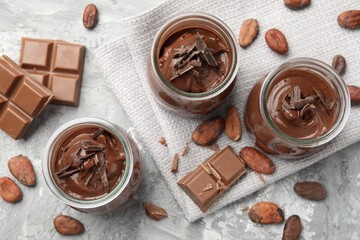 Tasty pudding in glass jars, pieces of chocolate and cocoa beans on light grey table, flat lay
