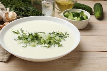 Tasty cucumber soup in bowl and ingredients on light wooden table, closeup
