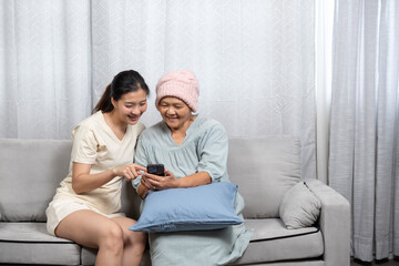 Obraz premium Young Asian woman helping middle-aged Asian female cancer patient wearing pink knitted hat use smartphone while sitting together on sofa, sharing joy, digital learning, emotional bonding, and support.