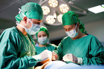 Medical students stitch artificial skin model, focusing on precision under intense surgical lighting