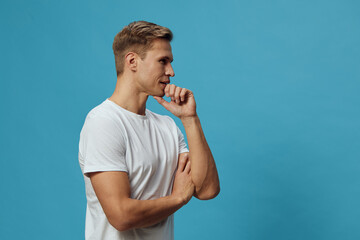 Smiling man with casual white t-shirt poses against a solid blue background, looking thoughtful and relaxed, isolated on plain background. People lifestyle concept