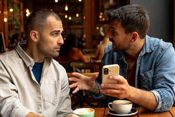 Two male friends discussing and arguing at a cafe, intently holding and examining a smartphone while sipping coffee together
