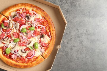Tasty pizza with smoked sausages and mushrooms in cardboard box on gray textured table, top view. Space for text