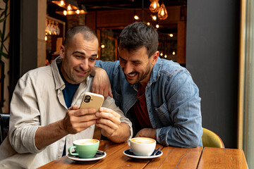 Two male friends enjoying a coffee break, looking at smartphone and laughing together at a cafe