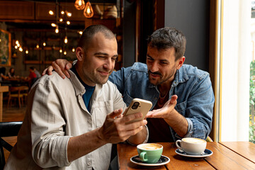 Two male friends enjoying coffee together at a cafe, happily browsing their smartphones and sharing...