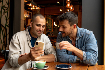 Two male friends sitting together at a cafe table, enjoying coffee while sharing moments on their smartphones and engaging in conversation