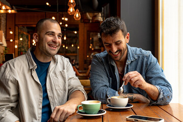Two male friends enjoying a cheerful coffee break, laughing and chatting while relaxing in a cozy cafe filled with warm ambiance