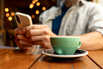 Hands holding a smartphone while enjoying a cappuccino on a wooden table in a cozy cafe, surrounded by softly blurred background lights