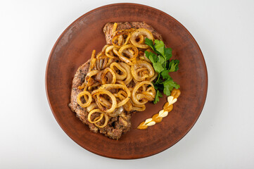 Grilled pork steak with golden fried onions served on a rustic clay plate. Traditional Eastern European dish with mustard, mayonnaise and fresh parsley garnish, on a light background