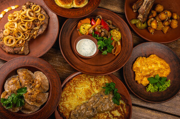 Traditional Eastern European cuisine served on rustic clay plates. Variety of homemade dishes including meat, potatoes, pancakes, dumplings, and grilled vegetables, on a wooden background