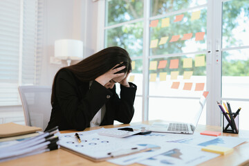 An overworked Asian businesswoman sits at her desk in a modern office, stressed and exhausted,...