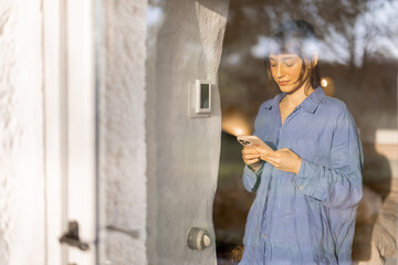 Woman in soft blue shirt holding smartphone, seen through a window at dusk, with reflections of...