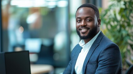 Fototapeta premium A smiling African American man in a suit and tie, seated at a desk in an office with a laptop and a green plant in the background.