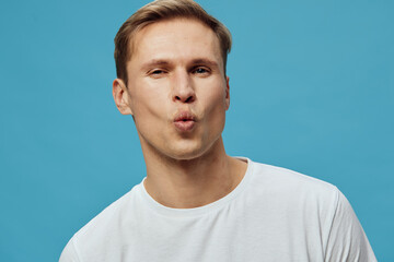 Fototapeta premium Confident young man with light brown hair making a playful kissy face, wearing a plain white t-shirt, shot against a bright blue background in a studio portrait, casual style, expression of fun and
