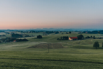 Obraz premium Idyllische Landschaft im West-Allgäu mit verstreuten Bauernhöfen, grünen Wiesen und majestätischen Bergen im Hintergrund – ein harmonisches Panorama voller natürlicher Schönheit vor Sonnenaufgang.