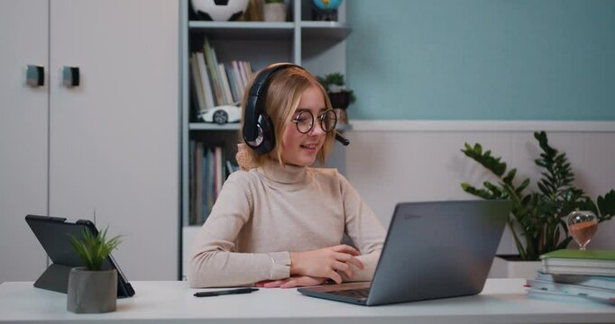 Smart caucasian kid girl wearing headphones waving hand talking with remote web teacher on social distance video conference call eLearning class on computer in living room at home