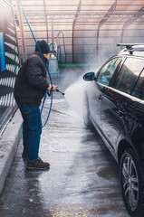 A man uses a high-pressure water sprayer to wash a black car at a car wash.