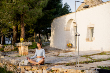Fototapeta premium A woman practices yoga in front of a traditional trullo in southern Italy. In a balancing pose, she connects with nature and morning light, grounding herself on ancient stone