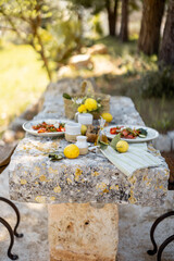A Mediterranean meal is served on a weathered stone table under the trees with lemons and olive...