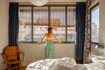 A woman stands by the window bathed in morning light, embracing a new day in her cozy Roman...