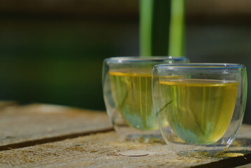 Two small transparent glass cups of green herbal tea placed on natural wooden surface outdoors ona sunny morning