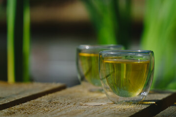 Two small transparent glass cups of green herbal tea placed on natural wooden surface outdoors ona sunny morning