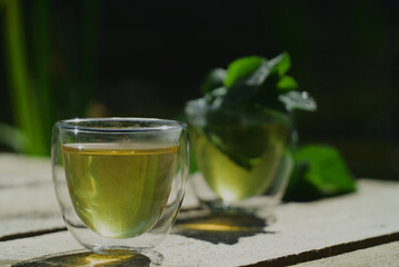 Two small transparent glass cups of green herbal tea placed on natural wooden surface outdoors ona sunny morning
