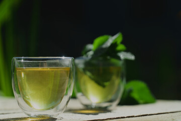 Two small transparent glass cups of green herbal tea placed on natural wooden surface outdoors ona sunny morning