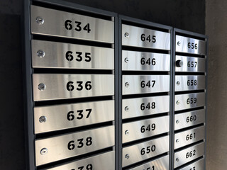 Row of modern stainless steel mailboxes with bold black numbers and built in locks mounted on dark wall in apartment building lobby, illustrating secure personal storage in urban residential space