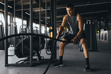 Full size body length of athletic man training with heavy battle ropes in a well equipped gym,...