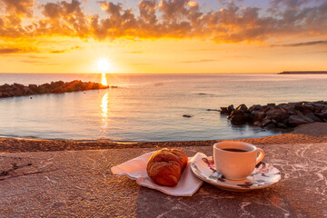 cup of coffee or tea with croissant on a morning embarkment beach with blue sea and beautiful cloudy sunrise or sunset on background, street food breakfast concept