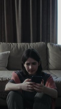 Medium full vertical of teenage Caucasian girl sitting hunched up on floor at home, finding vile content on social media, staring with disgust, sighing then turning away