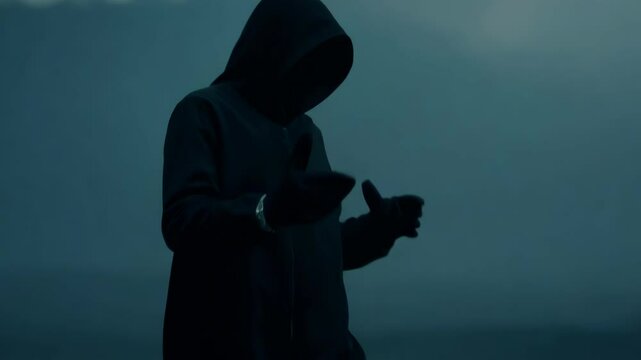 Mysterious hooded figure in silhouette gesturing expressively. Person moves hands dramatically while speaking or performing, creating powerful shadowy movements that convey intensity.