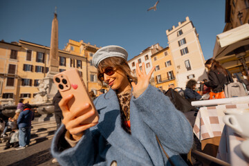 Woman in a blue coat scrolls her phone with a smile at a café table in a vibrant Roman square,...