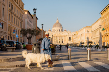 A woman in a blue coat walks her fluffy white dog across a Roman street with Saint Peters Basilica...