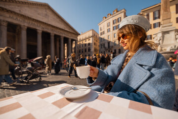 A woman in a blue coat enjoys her morning coffee at a table in front of the Pantheon in Rome,...