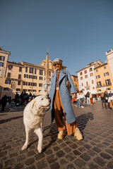 Stylish woman in a blue coat walking her fluffy white dog across a sunny Roman piazza filled with locals and tourists